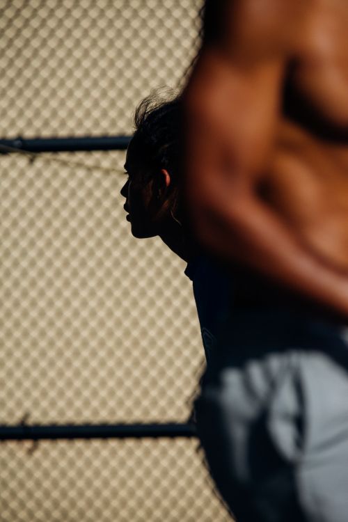 Profile of a person in silhouette behind the muscular torso of another person near a chain link fence.