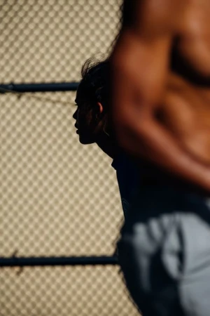 Profile of a person in silhouette behind the muscular torso of another person near a chain link fence.