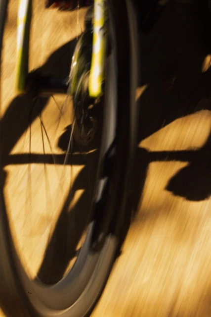 Close-up of a bicycle wheel in motion, displaying spokes and tire tread, set against a sunlit wood-grain background with shadows.
