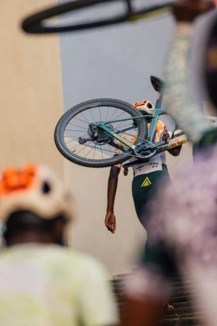 Person carrying a bicycle on their shoulder while walking up stairs, with another person in the foreground wearing a helmet.