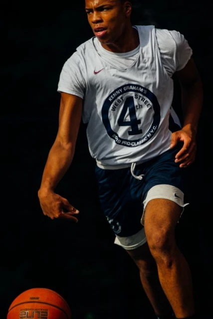 Action shot of an adult male basketball player dribbling a ball on an outdoor court with a dark background.