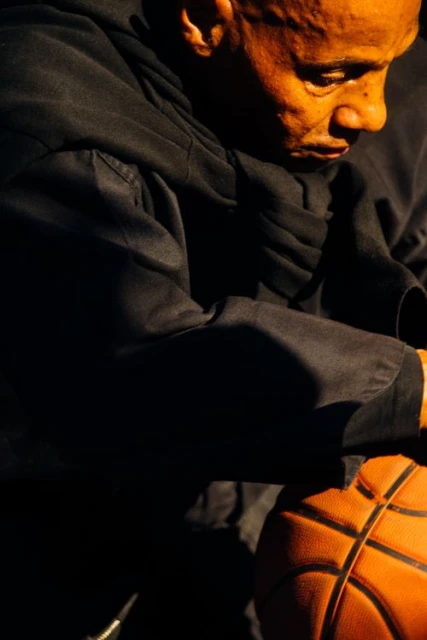 Close-up of a man in a hoodie holding a basketball, highlighting textures and warm lighting in an indoor setting.