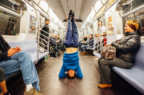 Person doing a headstand in a subway car with other passengers seated on either side of the train, wearing casual clothing.