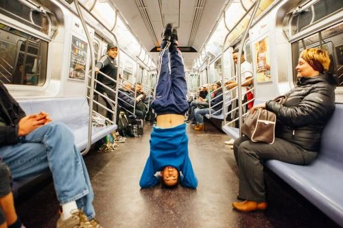 Person doing a headstand in a subway car with other passengers seated on either side of the train, wearing casual clothing.
