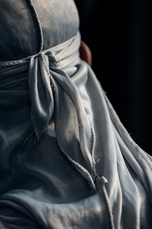Close-up of a person with draped grey fabric covering head, tied at the nape of the neck, with a dark background, and soft, diffused light.
