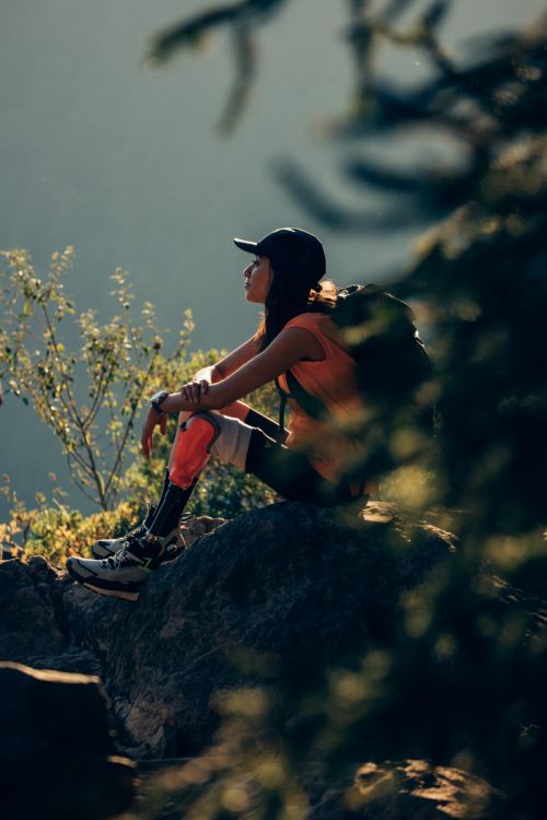 Woman with a backpack sits on a rock in an outdoor mountain landscape setting, wearing athletic gear and a baseball cap, looking contemplative.