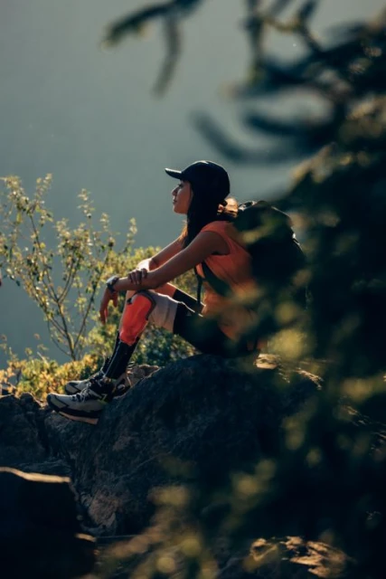 Woman with a backpack sits on a rock in an outdoor mountain landscape setting, wearing athletic gear and a baseball cap, looking contemplative.