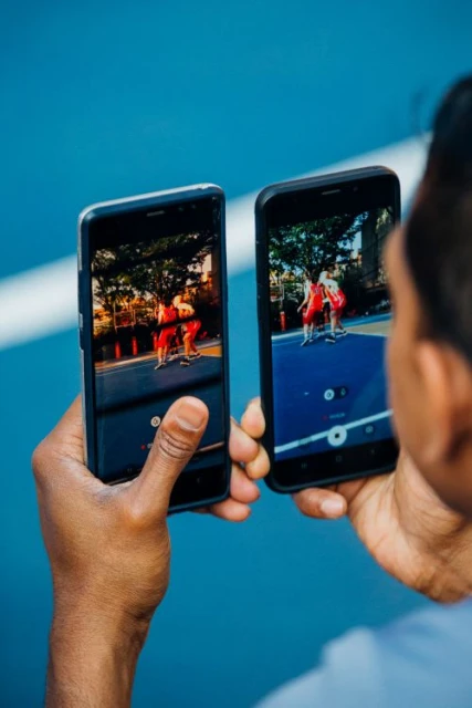 Person holding two smartphones displaying basketball players. Focus on mobile devices showcasing the game, captured in bright daylight.
