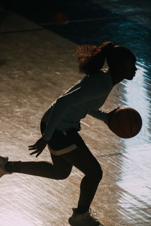 Silhouette of a woman dribbling a basketball on an indoor court, illuminated by a light source behind her, creating a strong contrast.