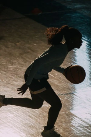 Silhouette of a woman dribbling a basketball on an indoor court, illuminated by a light source behind her, creating a strong contrast.