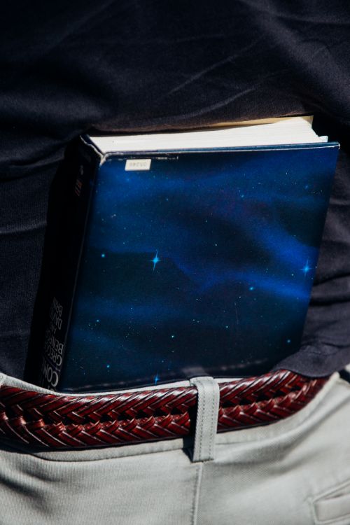 Close-up of a book with a space-themed cover tucked into the waistband of a person's light grey pants, secured with a braided brown belt.