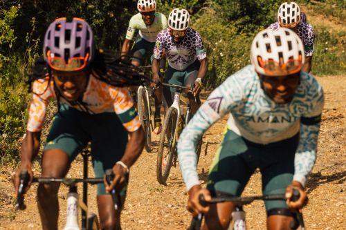 Group of cyclists in athletic gear compete in a race on a dirt path surrounded by green vegetation on a sunny day, captured at eye-level.