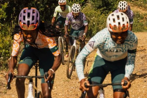 Group of cyclists in athletic gear compete in a race on a dirt path surrounded by green vegetation on a sunny day, captured at eye-level.