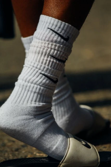 Close-up of an adult with dark skin wearing white Nike socks and slide sandals, set against a neutral outdoor background.