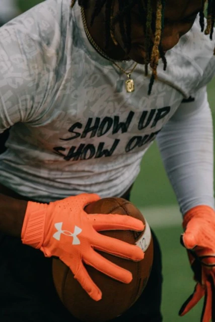 Football player in bright orange gloves holds a football, wearing a printed long-sleeve shirt and gold chain necklace. Indoor setting.