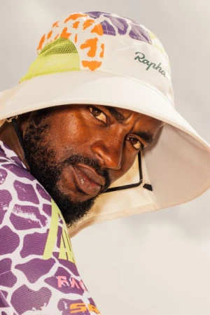 Black man wearing a patterned bucket hat and colorful athletic jersey, photographed in New York.