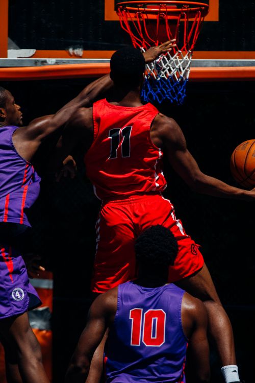 A group of basketball players compete during a game; one player is in red at the basket, and two other players are in purple uniforms.