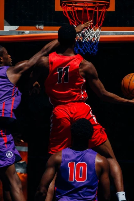 A group of basketball players compete during a game; one player is in red at the basket, and two other players are in purple uniforms.