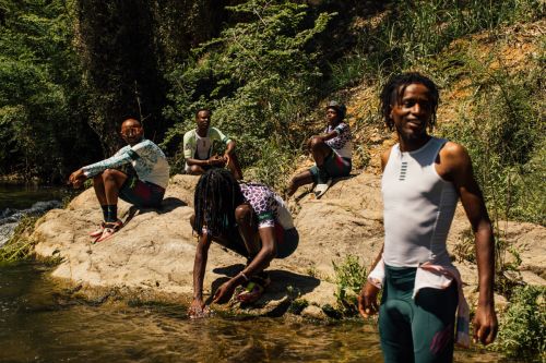 Group of young Black adults gather by a river on rocks in daylight. Some are partially submerged, surrounded by lush green foliage.
