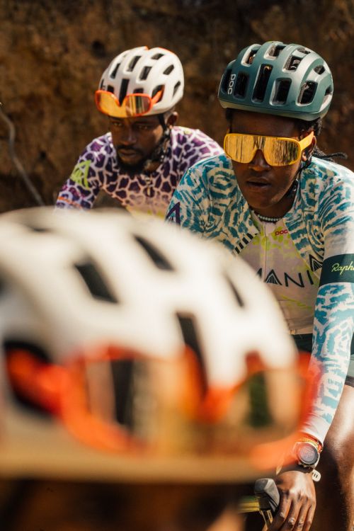 Two cyclists wear helmets and sunglasses while riding on a dirt path, with another cyclist's helmet visible in the foreground.