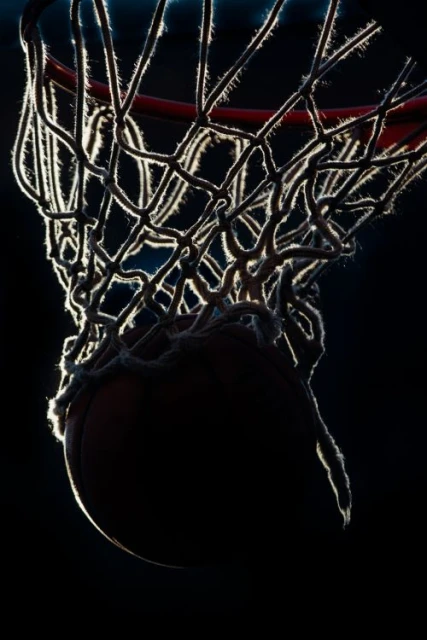 A silhouetted basketball passes through a net against a dark background, with the net fibers brightly illuminated by backlight.