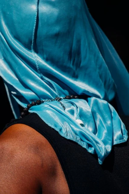 Close-up of an adult wearing a teal-colored silky hood or head covering, a black tank top, and a black beaded necklace with a silver cross.