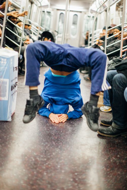 A young adult male in blue pants and hoodie balances upside down with hands on subway floor, with blurry commuters in the background.