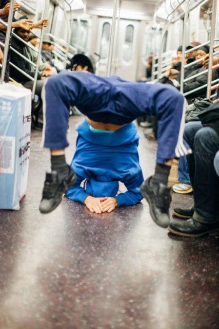 A young adult male in blue pants and hoodie balances upside down with hands on subway floor, with blurry commuters in the background.