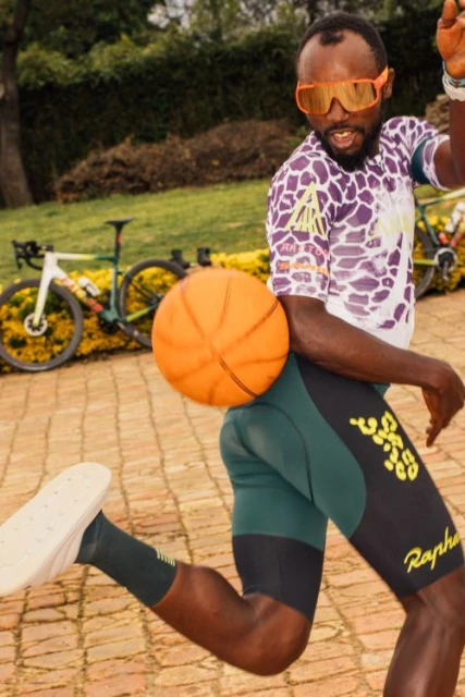 Adult male athlete dribbling a basketball in an outdoor setting, wearing sport clothing and orange sunglasses with a bicycle in the background.