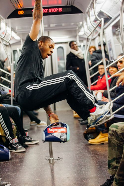 Man hanging from a pole in a subway car, wearing black Adidas tracksuit, sneakers, red socks, holding a boombox, passengers in background.