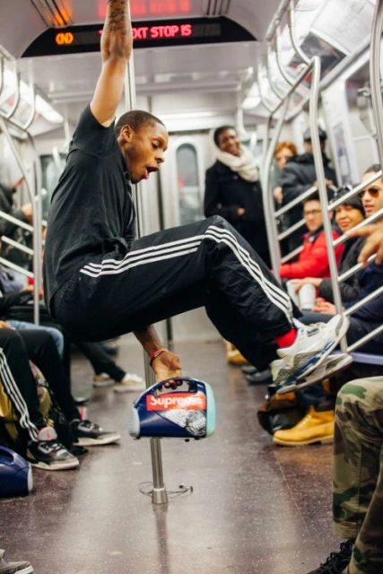 Man hanging from a pole in a subway car, wearing black Adidas tracksuit, sneakers, red socks, holding a boombox, passengers in background.
