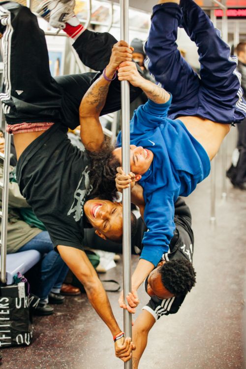 Three athletic young men perform acrobatic stunts on a subway pole, showcasing flexibility and balance in an urban transit setting.