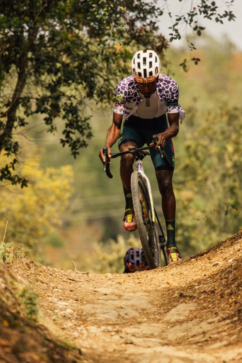 African-American male cyclist on a dirt path. Cyclist wears white helmet and colorful jersey, framed by trees and autumn foliage.