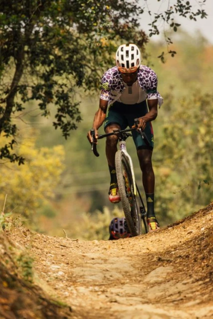 African-American male cyclist on a dirt path. Cyclist wears white helmet and colorful jersey, framed by trees and autumn foliage.