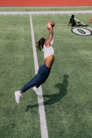 African American man makes a diving catch with a football on green football field in the daytime with other people in the background.