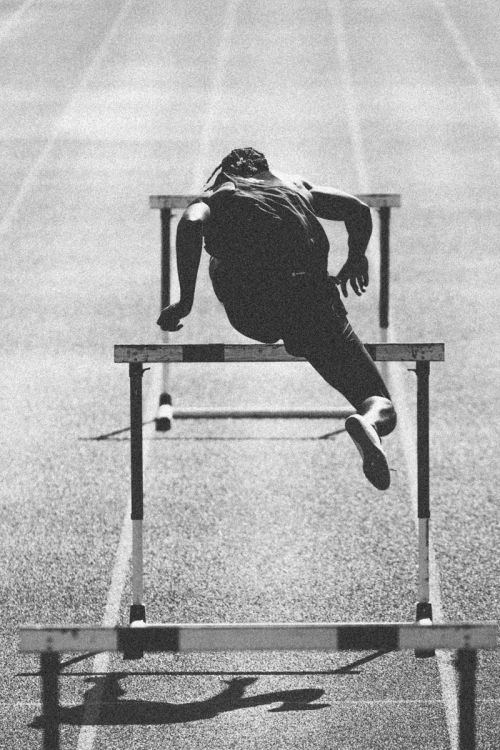 Monochrome action shot of a muscular athlete clearing a hurdle on a running track, showcasing speed and athleticism in a sports competition.