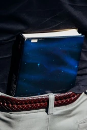 Close-up of a book tucked into the back waistband of a person wearing light trousers and a woven brown leather belt.