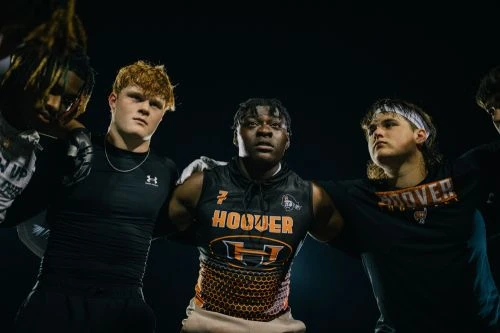 Low-angle shot of a group of male football players in black athletic jerseys with arms around each other in a huddle at night.