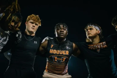 Low-angle shot of a group of male football players in black athletic jerseys with arms around each other in a huddle at night.