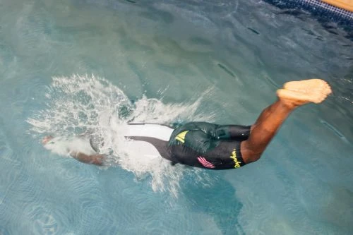 Swimmer diving into an outdoor swimming pool, captured mid-motion with splashing water around the head and shoulders.