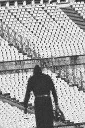 Monochrome rear view of a hooded person standing before rows of empty stadium seats by photographer Alessandro Simonetti