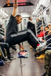 Young man swinging on a subway pole wearing a black t-shirt and black track pants with white stripes, photographed by Alessandro Simonetti in New York.