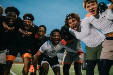 Group of young male athletes in athletic wear shouting and celebrating together in a huddle on a football field.