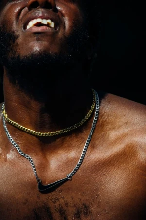 Close-up portrait of a shirtless Black man wearing a gold chain and a silver chain with a Nike pendant against a dark background