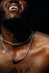 Close-up portrait of a shirtless Black man wearing a gold chain and a silver chain with a Nike pendant against a dark background