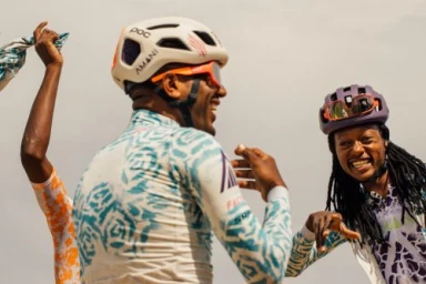 Three cyclists in patterned jerseys and helmets smiling outdoors under an overcast sky, photographed by Alessandro Simonetti.
