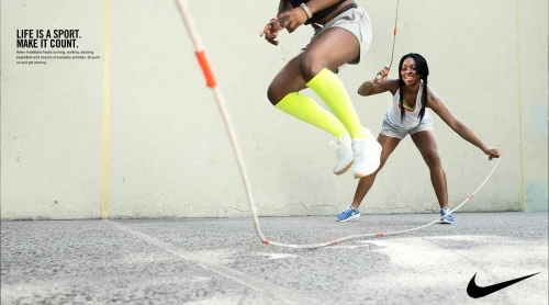 Two Black women jumping rope outdoors on pavement wearing athletic clothing and smiling, promoting sport and fitness.