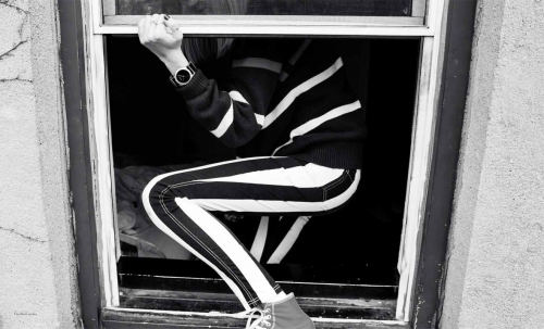 Black and white portrait of a person crouching inside a window frame wearing striped pants and holding the frame ledge