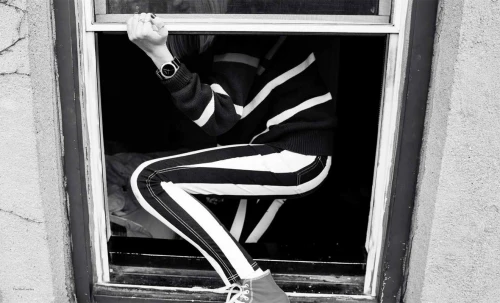 Black and white portrait of a person crouching inside a window frame wearing striped pants and holding the frame ledge