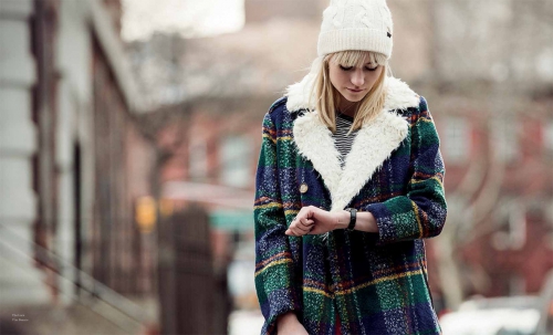 Young woman in a patterned coat and white knit hat checks her watch outdoors against a blurred urban street background.  